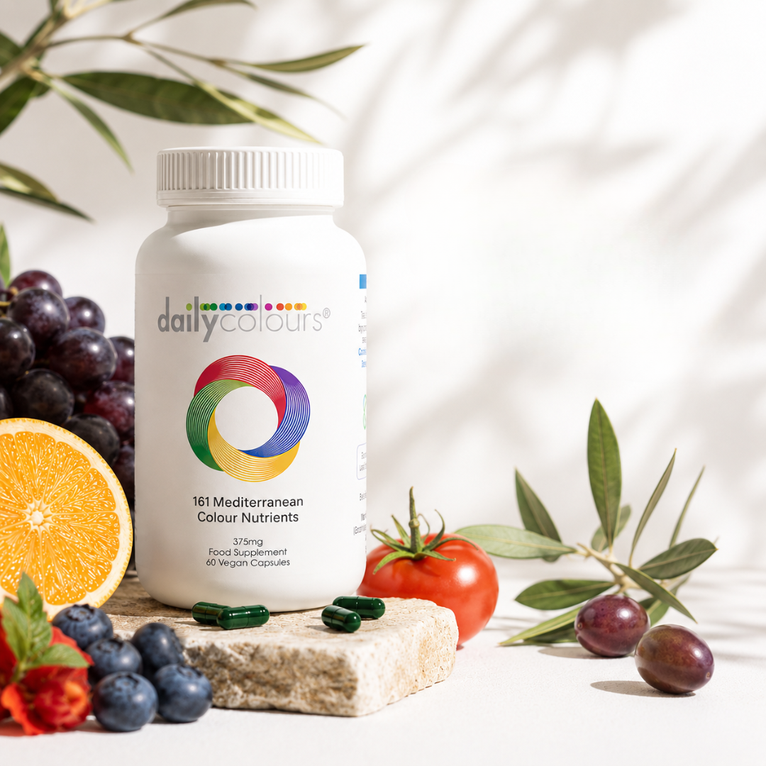 A white bottle of dailycolours 181 Mediterranean Colour Nutrients food supplement stands on a stone slab surrounded by fresh fruits and vegetables, including grapes, an orange slice, blueberries, a tomato, and olives, with leafy green shadows on a white background.
