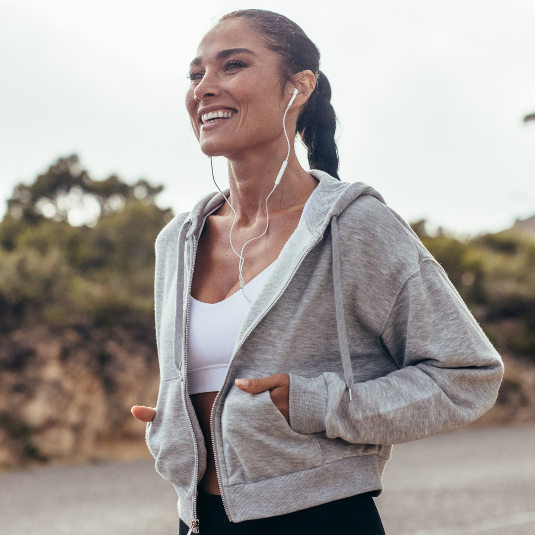 A woman smiling outdoors while wearing a light gray zip-up hoodie over a white sports bra and black leggings, with white wired earphones in her hair.